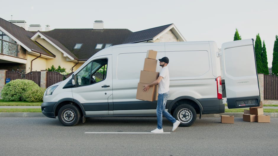 A man wearing a white t-shirt, light blue jeans, white sneakers, and a black cap is engaged in a home relocation task, carrying three cardboard boxes of varying sizes while standing on the street in front of a white delivery van. The van, branded for a removals service, is parked parallel to the curb with its rear doors open, revealing an empty cargo area prepared for furniture and packing materials. Additional cardboard boxes are placed on the pavement next to the van, indicating an active packing and loading process. The background features residential houses with dark roofs, large windows, and fencing, alongside well-maintained bushes and trees, under daytime lighting conditions. The scene is centered on the transportation phase of furniture transport and packing and moving activities, with the man actively loading or unloading household items as part of a professional removals operation, exemplified by companies such as Man With a Van Brent.