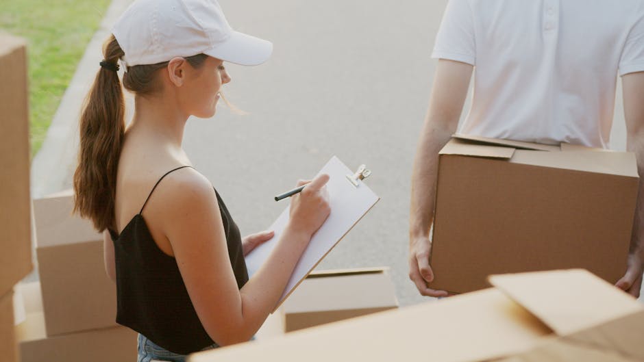A young woman with long brown hair tied in a ponytail wearing a white cap and a black sleeveless top is standing outdoors on a paved surface, next to a stack of cardboard boxes, and is holding a clipboard with a pen to take notes. She is facing a man dressed in a white t-shirt, who is holding a medium-sized brown cardboard box. The background shows a portion of a building or property entryway, and the scene is well-lit with natural daylight. The setting appears to be part of a home relocation or moving process, with packing and logistics preparations visible, including the boxes and the outdoor environment indicating the loading or unloading stage as part of the services offered by Man With a Van Brent, specializing in removals.