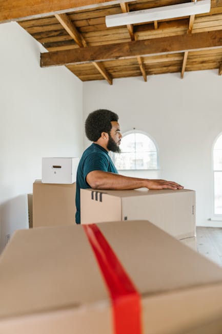 Interior image depicting a light-filled room with a light wooden table against a white wall, supporting several cardboard boxes labeled 'KITCHEN' and 'OFFICE', some wrapped in protective plastic or pa