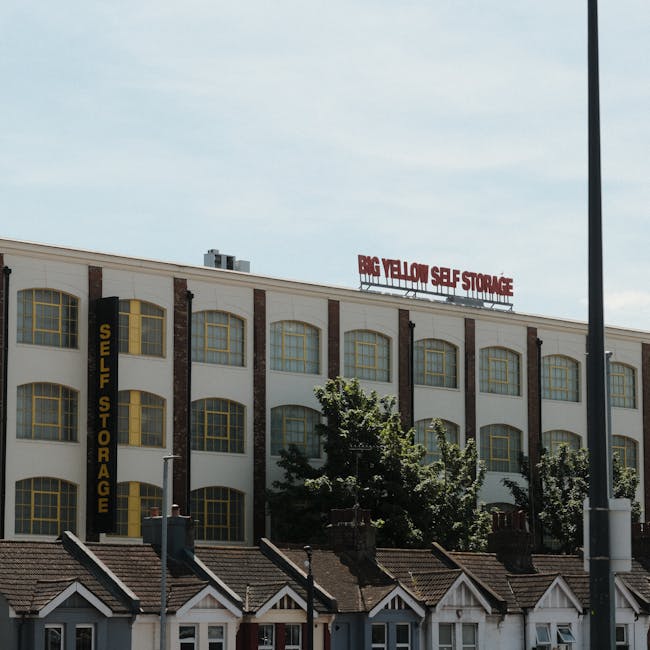 A multi-storey building with a white facade and large yellow-framed windows, featuring signage indicating 'Self Storage' and 'Big Yellow Self Storage' on the roof. The building is situated behind a row of residential houses with tiled roofs and small chimneys. In front, there are several trees, a lamppost on the right side, and a paved pavement area. The scene is well-lit with natural daylight, and the overall setting suggests a commercial storage facility, relevant to home relocation and furniture transport services offered by Man With a Van Brent, as seen in the context of house removals and packing and moving logistics.